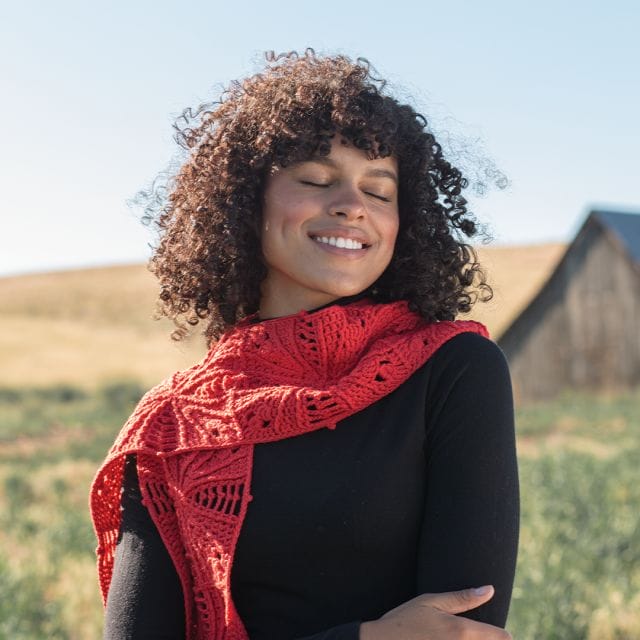 A woman stands in the High Desert of Oregon, wearing a hand-crocheted scarf made in red.