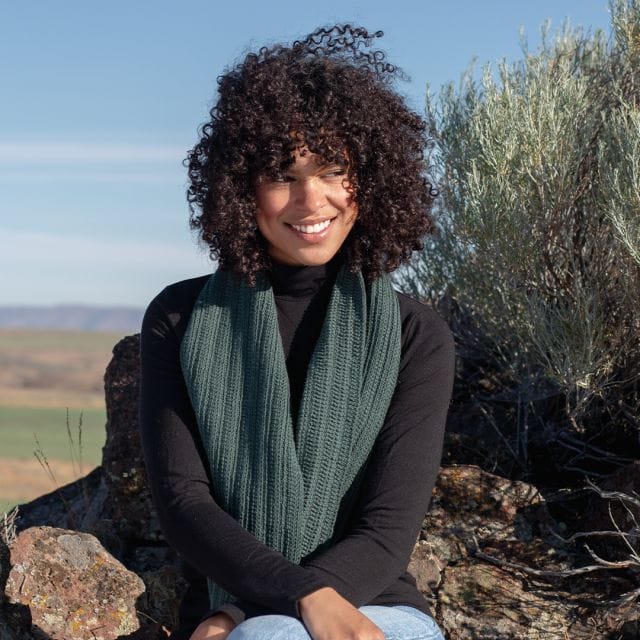 A woman sits in Oregon's High Desert wearing the Willowdale Tunisian Scarf, a crochet pattern available from crochet.com