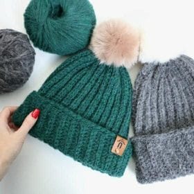 A hand touching a green crocheted beanie with a tan fur pom-pom. Nearby, a gray beanie with a white pom, and 2 balls of yarn in gray and green.