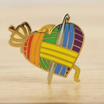An enamel pin that is shaped like a heart with rainbow colored yarn wrapped around it, a crown, and a crochet hook