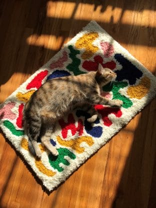 A cat laying on a tufted rug with abstract colorful shapes on a white background