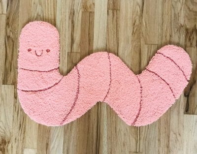 A punch needle/tufted rug shaped like a wiggly pink worm with a smile