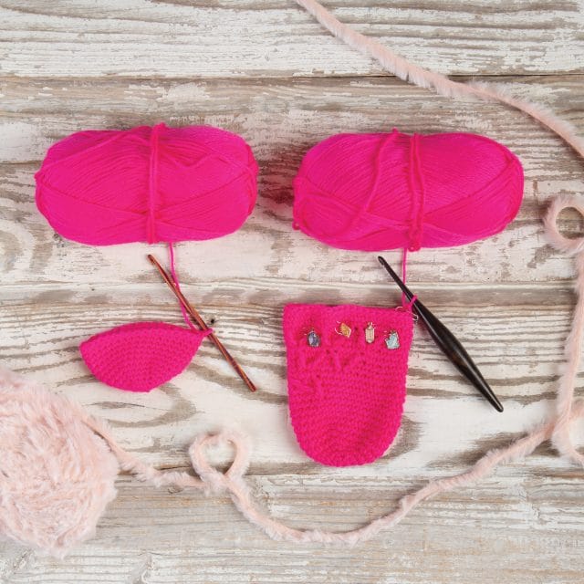 On a board background, two partially-finished Summit Slipper Socks in hot pink, along with hooks and yarn balls
