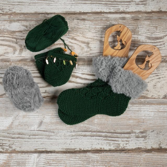 On a board background, a pair of finished summit slipper socks on sock blockers, along with a partially-finished pair and a couple balls of yarn